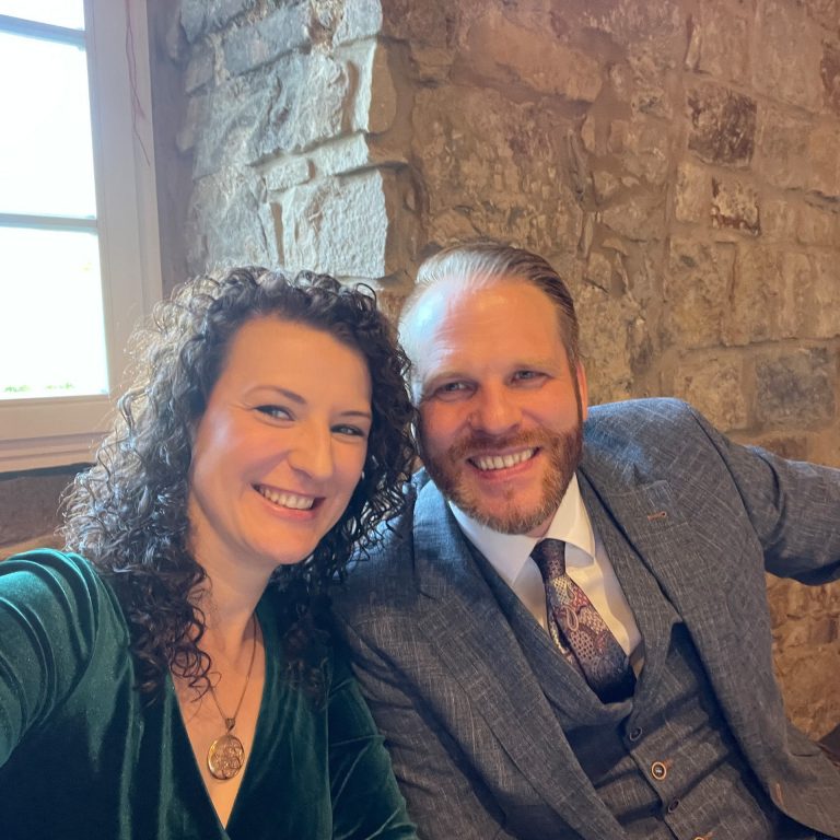 Ein Paar lächelt in die Kamera mit steiniger Wand im Hintergrund auf einem festlichen Anlass , Cozyhomes Ferienimmobilien, Ferienwohnung Bad Arolsen, Ferienhaus Kellerwald, Ferienhaus Edersee, Ferienwohnung Arolsen